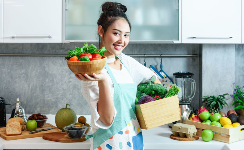 mujer en la cocina sosteniendo varios vegetales y frutas en sus brazos